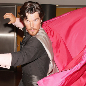 Benedict Cumberbatch-inspired hero, striking pose with dark wavy hair and close-cut beard. Dressed in dark outfit with vibrant red cape extension. Blurred background, dramatic lighting effect.