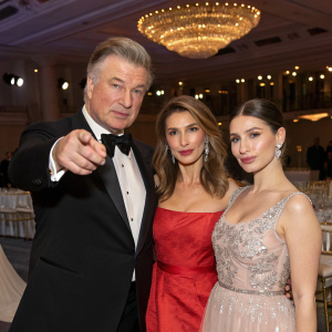 Family portrait: Alec Baldwin, Hilaria Baldwin, and Carmen Gabriela Baldwin at a gala event. Alec Baldwin is pointing towards the camera. The setting is a formal hall with chandeliers.