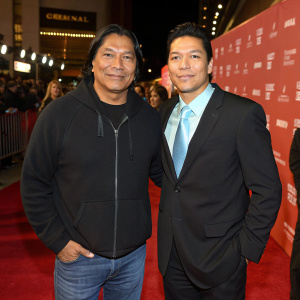 Gil Birmingham and Chaske Spencer at the Sundance Film Festival. Gil is in a black hoodie and jeans. Chaske is in a dark suit with a light blue tie. Both are Native American. Against a bustling red carpet backdrop.