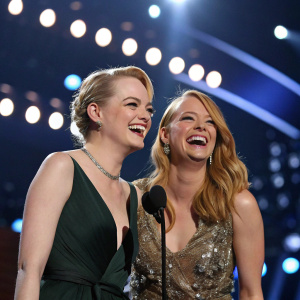 Emma Stone and Krista Stone laughing together at the Grammy Awards. The stage background is filled with spotlights.