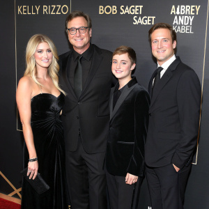 At a glitzy Hollywood gala, Kelly Rizzo, Bob Saget, Aubrey Saget, and Andy Kabel strike a pose. Kelly in a stunning black velvet gown, Aubrey in an elegant black dress, while Andy and Bob don matching dark suits. The backdrop is a sleek black with minimalistic gold text and logos.