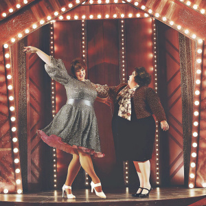 Tracy Turnblad and Edna Turnblad performing on an elaborate Broadway stage. Vibrant costumes and colorful set design
