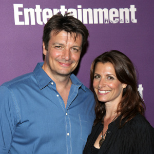 Nathan Fillion and Darla Delgado, smiling and posing together at the Entertainment Weekly and Syfy party. Nathan in a blue button-up shirt, Darla in a dark top. Solid purple background with large 'Entertainment' text.
