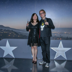 Jennifer Tilly and Phil Laak at the Hollywood Walk of Fame unveiling. Jennifer in a black sequined dress, Phil in a black jacket with a cartoon graphic. They pose with champagne glasses, against a backdrop of stars and city lights.