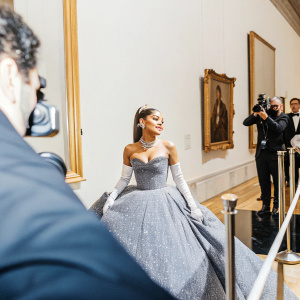 Ariana Grande at the gala event. Wearing a glittery, off-the-shoulder gown. Surrounded by flashing cameras and guests. Museum setting.
