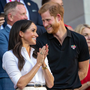 Meghan Markle, smiling and clapping in a white blouse, stands beside Prince Harry, who is dressed in a black polo shirt. They are at a charity event surrounded by other guests.