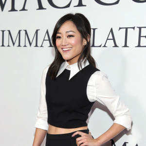 Karen Fukuhara, actress, posing in a black crop top with a white shirt underneath. The backdrop is white with black text. Immaculate lighting highlights her cheerful smile and sleek, straight hair.