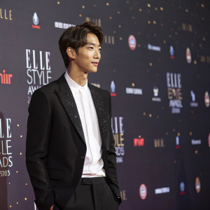 Kim Young-Kwang at Elle Style Awards. Wearing a black suit and white shirt. Against a dark backdrop filled with flashing lights and logos.