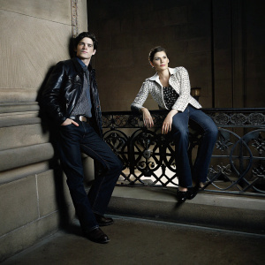 Caucasian actors Billy Crudup and Maggie Gyllenhaal, stylishly dressed, posing for a magazine cover shoot. Billy in black leather jacket, casual shirt, and pants. Maggie in a checked jacket and blouse with dark jeans. Setting: vintage New York street scene with elaborate ironwork railing.