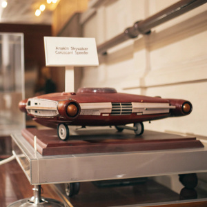 A detailed exhibit showcasing Anakin Skywalker's Coruscant Speeder from Star Wars. The speeder is displayed on a wheeled platform inside a museum. The platform is set on a structured floor with a white sign providing information about the speeder's significance and design.