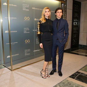 Nicola Peltz in a sophisticated black blouse and pencil skirt, paired with strappy heels. Will Peltz dons a navy suit. They are at a glamorous awards ceremony, standing behind a sleek, glass wall adorned with corporate branding.