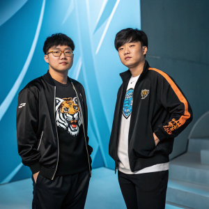 Heo 'ShowMaker' Su and Jeong 'Chovy' Ji-hoon, Korean League of Legends pros. ShowMaker in tiger graphic t-shirt. Chovy in jacket and athletic wear. Posing in studio with abstract blue backdrop.
