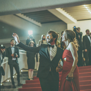 Gaspard Ulliel and Gaelle Pietri at a glamorous film festival premiere. They are dressed in exquisite evening wear. The background features bustling crowds and flashing cameras.