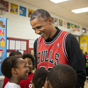 Barack Obama, wearing a Chicago Bulls jersey, surrounded by a group of young children at a community center event.