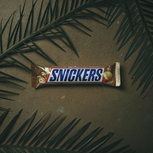 Modern design of Snickers chocolate bar wrapper. Features Arabic script, sleek brown exterior, and white interior. Packaging shot