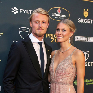 Simon Kjaer and Elina Gollert, a dapper Danish footballer and his elegant wife, posing for photographers at a glamorous awards ceremony. The backdrop features bright sponsor logos
