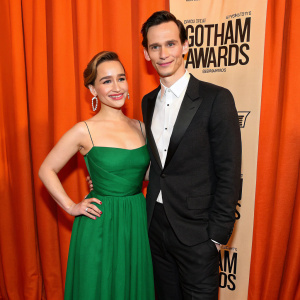 Emilia Clarke and Cory Michael Smith at the Gotham Awards. They are both dressed formally, Emilia in a green gown, Cory in black and white attire. The backdrop is a vibrant orange curtain.