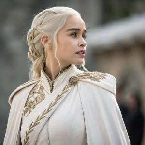 Close-up shot of Emilia Clarke as Daenerys Targaryen. She wears a dramatic, flowing white robe with gold embroidery. Intense, focused look