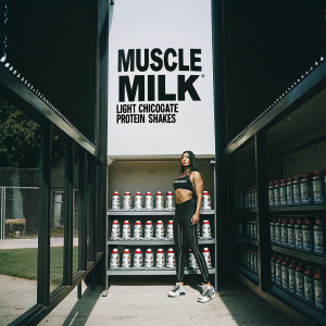 Promotional image for Muscle Milk Light Chocolate Protein Shakes. Shelves lined with various flavors and a woman in fitness gear