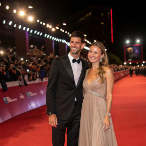Novak Djokovic and Jelena Ristic attending the star-studded premiere of a major film festival. Novak in black tuxedo, Jelena in long taupe gown. Red carpet illuminated with flashbulbs and surrounded by enthusiastic fans.