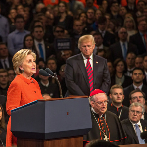 Hillary Clinton addressing a large audience. Donald Trump and Cardinal Dolan stand silently behind her. A packed auditorium with various political figures and supporters in attendance.