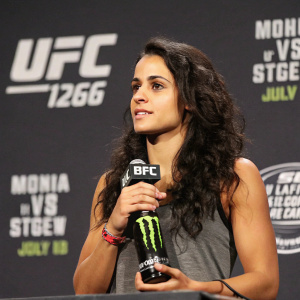 Ariane Lipski, MMA fighter, at the post-fight press conference. Dark wavy hair, casual attire, holding a Monster Energy water bottle. The backdrop features UFC branding.