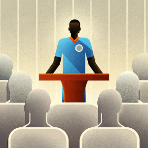 Riyad Mahrez, wearing a blue sports jersey with Manchester City logo. Attending a press conference, blurred audience in the background.