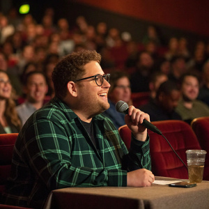 Jonah Hill, wearing a green plaid shirt and glasses, participating in a Q&A session at a packed theater.