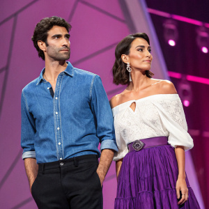 Fabio Mancini and Caterina Balivo on a glamorous event stage. Fabio in blue denim shirt and black pants, Caterina in white off-the-shoulder blouse and purple ruffled skirt. Vibrant purplish-pink backdrop. Serious expressions