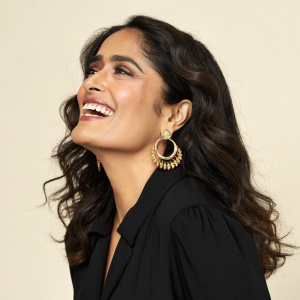 Salma Hayek, ethnic female, beaming smile, black blouse, large gold earrings. Wavy dark brown hair. Studio setting, plain beige backdrop.