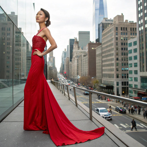 Akiko Chubachi, Asian model, posing in a fashion shoot. She wears a long red evening gown. New York Fashion Week, busy cityscape in background.