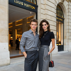 Xavier Samuel and Shermine Shahrivar at a luxury fashion event in Paris. Xavier in a stylish gray shirt and dark pants, Shermine in a black top and striped skirt. They pose confidently in front of a luxurious Louis Vuitton storefront.