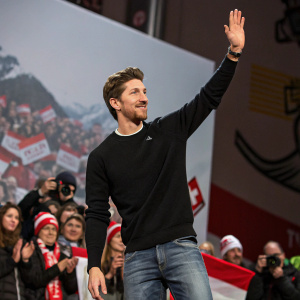 Marcel Hirscher, an Austrian skier, waving at the press conference. He is wearing a casual black sweater and jeans. The background is crowded with cheering fans.