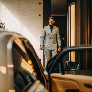 Bastian Schweinsteiger, in a sleek grey suit, standing at the entrance of a luxury car unveiling event. The backdrop is a sleek modern wall with subtle branding.