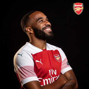 Alexandre Lacazette, with a red and white Arsenal jersey, smiling confidently. He has a short beard. Black background with sport brand logo
