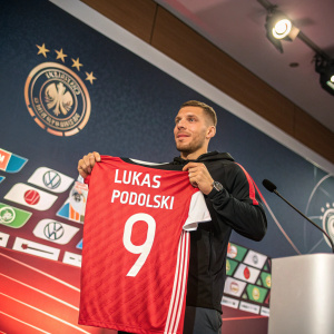 Lukas Podolski at a press event. Holding a jersey with his name and number. Backdrop has team logos and sponsor brands. Even lighting, vivid colors