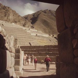 At an ancient Inca site in Peru, Imposing stone steps carved into the mountainside reveal the remarkable ingenuity of the Inca civilization. Tourists wander around, capturing the site's majesty with their cameras. The photograph, taken on a sunny day, highlights the intricate stone work and the towering mountain backdrop in vivid detail.