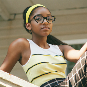 Marsai Martin, a young actress, in a vibrant yellow headband and black framed glasses, wearing a white tank top with horizontal stripes, her hair tied back in a ponytail. Posing for a formal event against a plain backdrop.