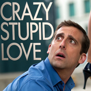Promotional image for 'Crazy, Stupid Love.' Close-up shot of Steve Carell in a blue shirt looking shocked. A woman's high-heeled foot and arm visible. Background blurred, focusing on the title 'CRAZY STUPID LOVE' in bold letters.