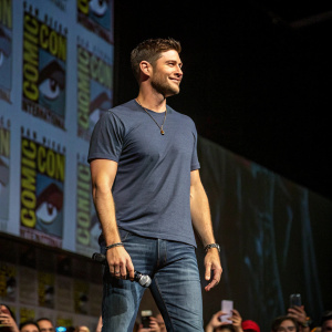 Jensen Ackles, wearing a casual t-shirt and jeans, appears on stage at a Comic-Con panel. A sea of excited fans fills the background.