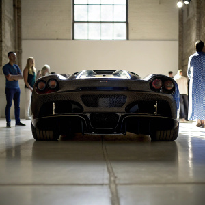 Exhibition photo of a black Ferrari LaFerrari Aperta. Rear view with white floor. Carbon fiber bodywork, exhausts. Attendees visible in ambient lighting.