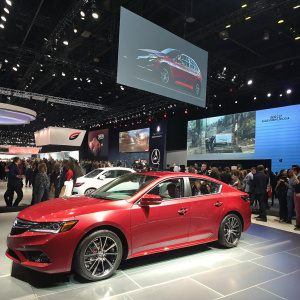 Shiny Acura ILX on display at car expo. Large screens showcasing features. Crowded event space.