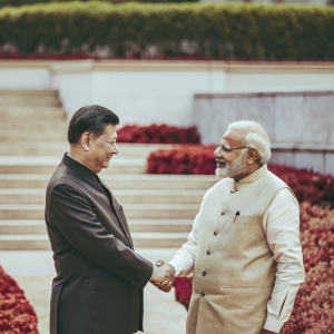 Xi Jinping and Narendra Modi in a friendly handshake during a state visit. Lush green gardens as the backdrop.