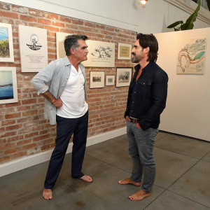 Peter Gallagher and Colin Farrell engaging in conversation at an art gallery opening. They are both barefoot and dressed in casual attire. The backdrop features a brick wall adorned with event branding and logos. The setting is informal with various art pieces displayed.