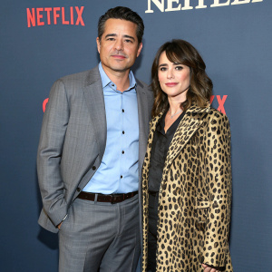 Steve Rodriguez and Linda Cardellini posing for a Netflix promotional photo. Steve in light blue shirt and grey suit, Linda in leopard print coat. Dark blue background with bold Netflix branding. Even lighting, modern tone.