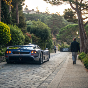 Maserati MC12 Corsa, parked on a serene, cobblestone street in Carmel-by-the-Sea, California. Lush greenery surrounds the car. Pedestrian walking by, admiring the angular rear wing and bold front end. License plate reads 'CORSA'.