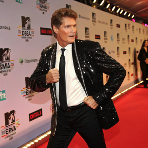 David Hasselhoff, at an MTV Europe Music Awards event, Berlin. Dressed in a black sequined jacket with a white shirt and black tie. Confident pose on red carpet with backdrop featuring sponsor logos like Sony Ericsson and Dell.