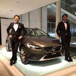 Tomas Ernberg and Sudeep Narayan, in tuxedos, stand beside the shiny Volvo V40 'Cross Country' at the gala unveiling. Sleek car showcased on a glass platform. Minimalist showroom with large windows and clean lines, setting the scene for the automotive event.