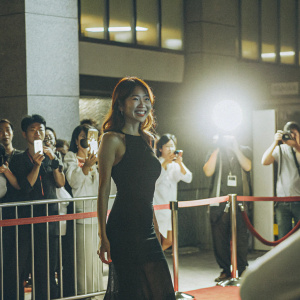 Seol In-ah, South Korean actress, posing confidently in a sleek black dress. A busy movie premiere scene with flashing cameras and audience in the background.