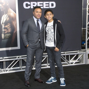 Oscar De La Hoya and Devon De La Hoya at the 'Creed' premiere. Oscar in a dark grey suit, tie and confident pose. Devon in graphic t-shirt and casual sneakers. Movie-themed backdrop with logo